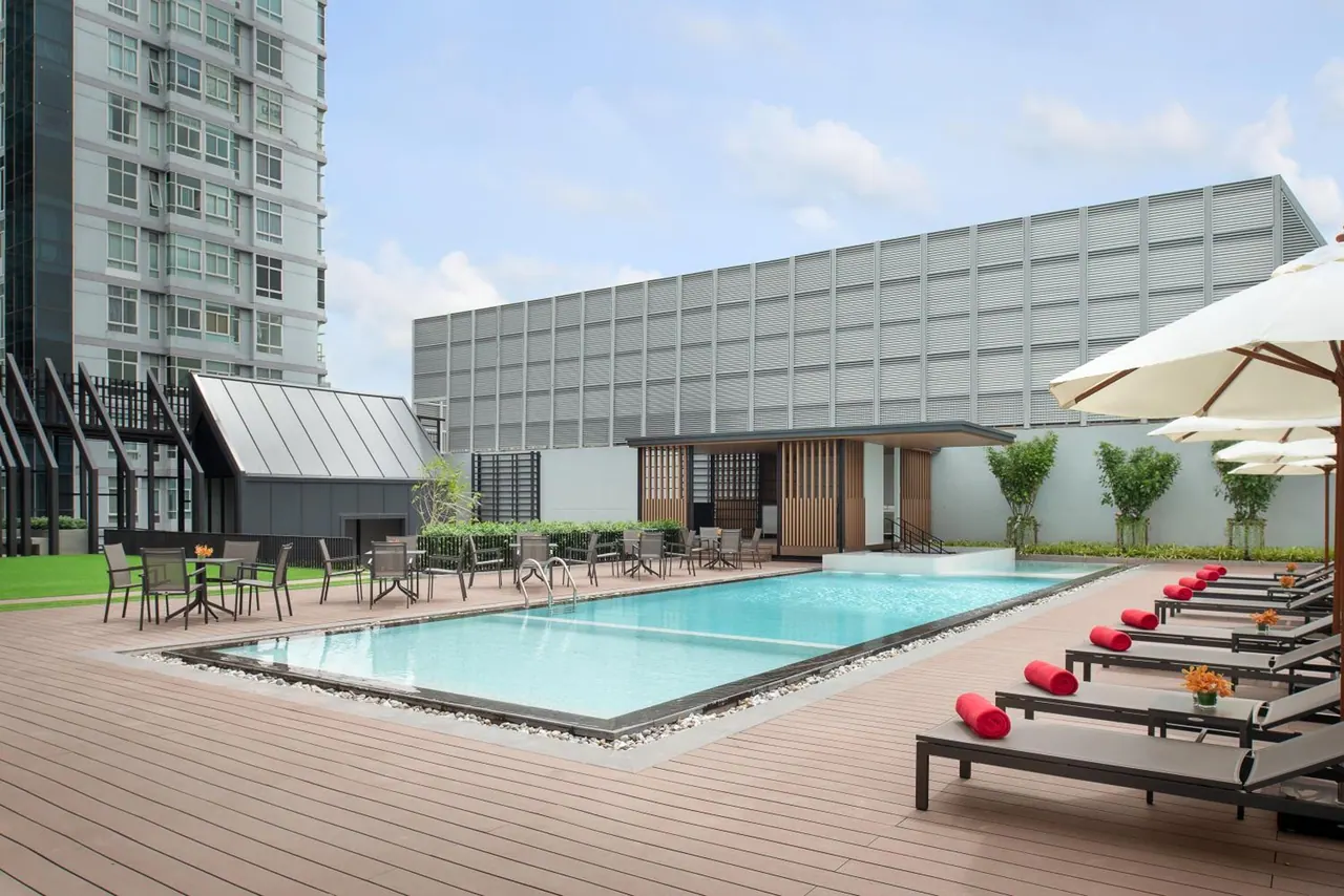 An outdoor rectangular swimming pool on a timber deck, with white umbrellas and red-bolstered sun loungers beside it and a tall residential tower on the left.
