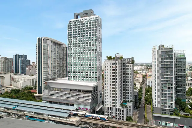 Elevated view of a tall hotel tower with an Avani sign on its roof, surrounded by other high-rise buildings, a street, and an elevated BTS train line below.