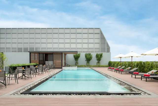 A rectangular rooftop swimming pool with wooden decking, red-cushioned sun loungers under cream parasols, and dining tables with grey chairs beside a louvred grey building.