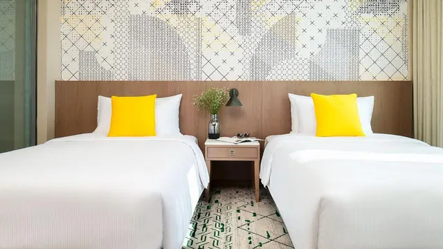 Two twin beds with white linen and yellow cushions sit against a wooden headboard beneath patterned wallpaper, with a bedside table between them.