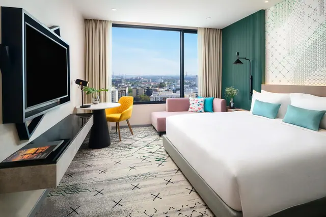 A hotel bedroom with a king bed, a pink sofa, a yellow chair at a round table, a wall-mounted television and a window overlooking a city.
