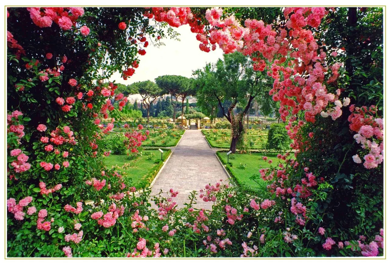 A paved path runs through a formal garden with lawns, trees and climbing pink roses framing the view.