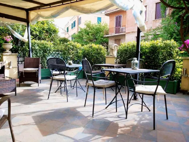 An outdoor terrace under a fabric canopy has metal tables and chairs, a wicker armchair, potted plants, and hedges, with a multi-storey building behind.