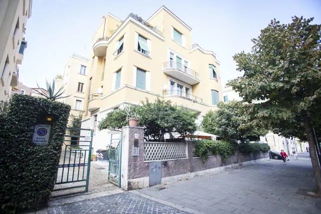 Exterior of a yellow multi-storey apartment building with balconies and green shutters, seen from a street with a gated entrance, a large tree and two pedestrians.