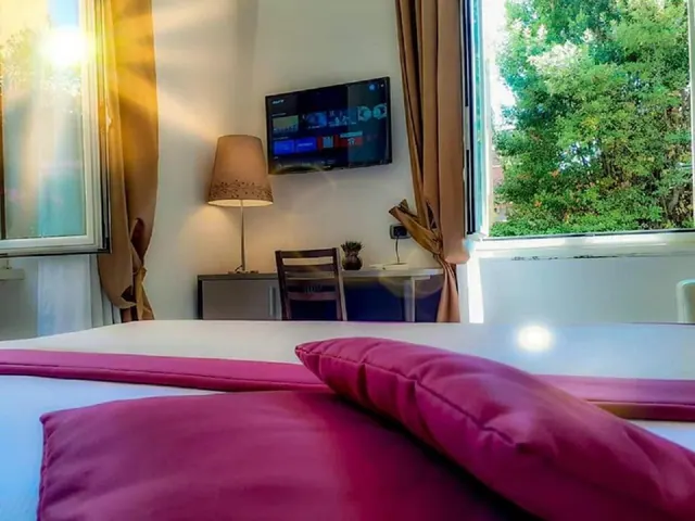 A bedroom with a made bed with magenta cushions in the foreground, a wall-mounted television, a table lamp, a desk with a chair, and a window showing green trees outside.