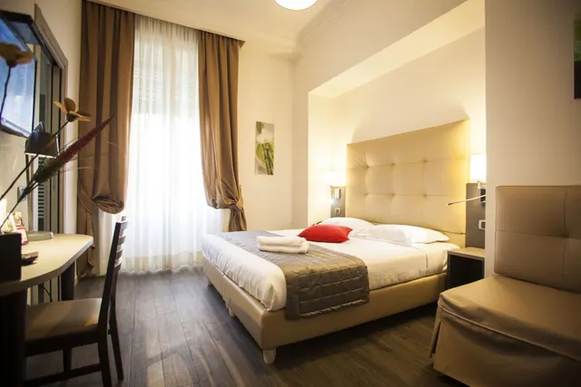 A double bed with a tufted headboard, a red cushion and folded towels sits in a bedroom beside a desk, an armchair and a window with sheer curtains.