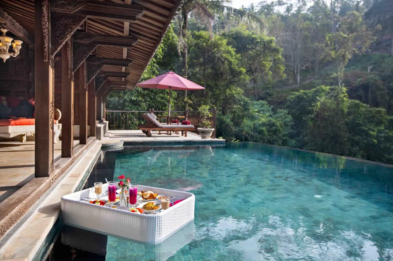 A breakfast tray with food and drinks floats in a pool beside a covered wooden pavilion, with jungle trees behind.