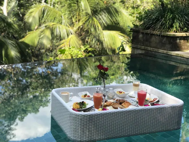A breakfast tray with plates, glasses and a rose in a vase floats on a pool, with palm trees and tropical greenery behind it.