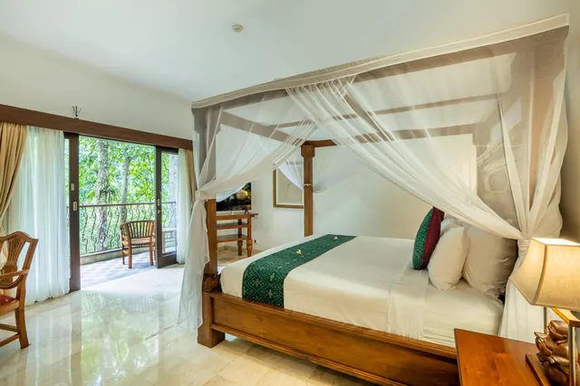A four-poster bed with a mosquito net stands in a bedroom, with glass doors open onto a balcony overlooking green trees.