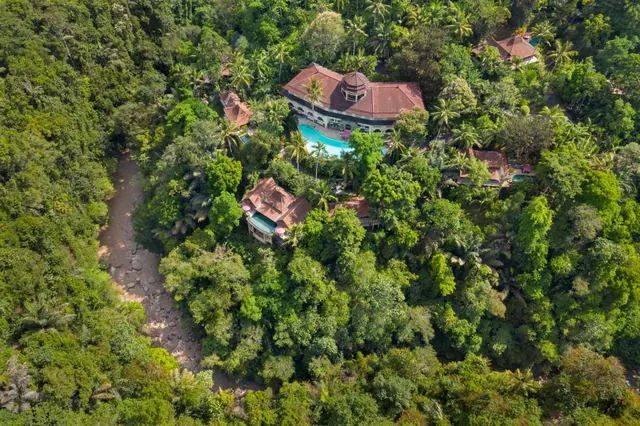 Aerial view of a red-roofed resort building with a pool and separate villas surrounded by dense jungle and a dry riverbed.