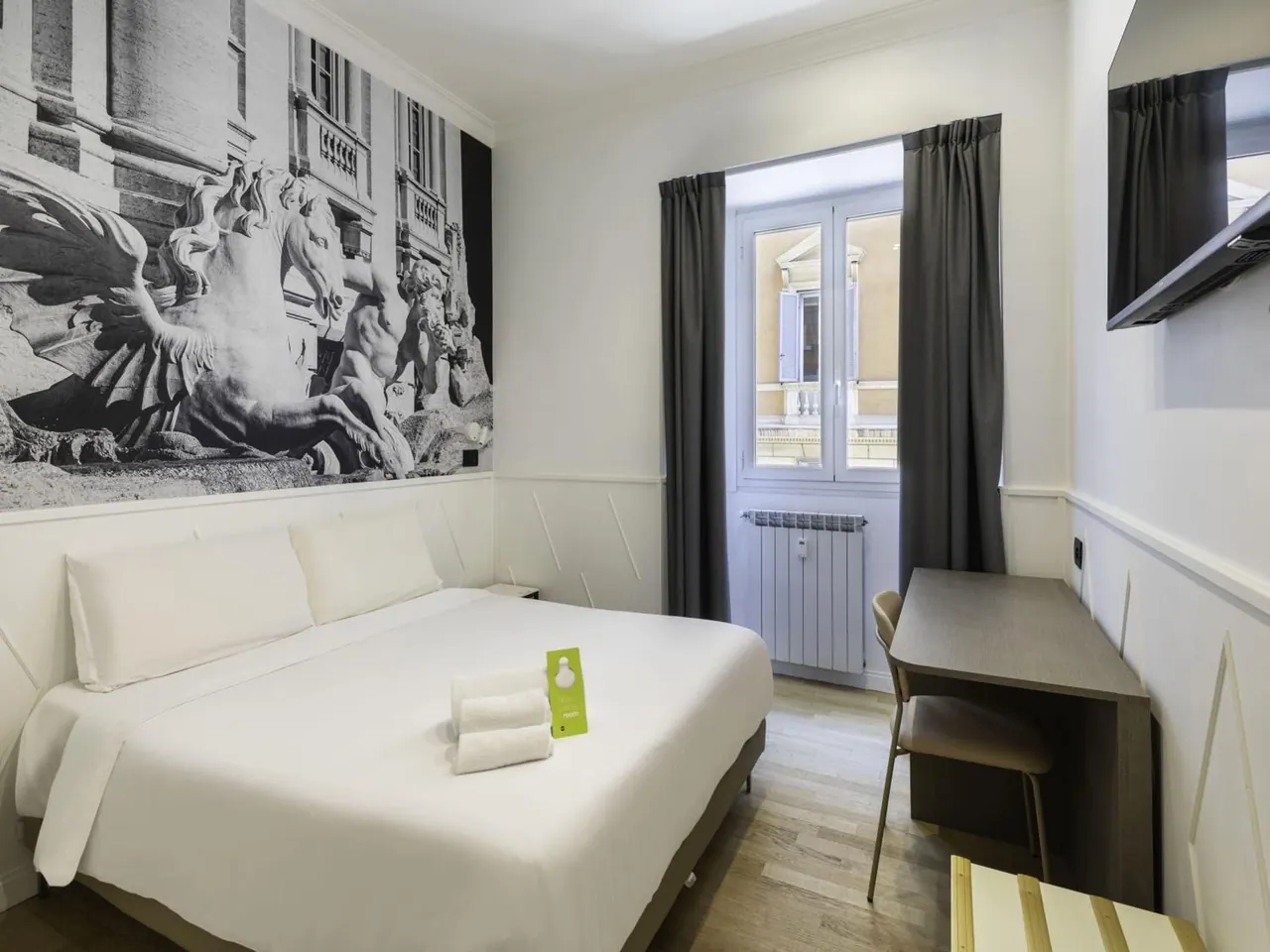 A bedroom with a double bed, a black-and-white sculpture mural on the wall, a desk with a chair, a wall-mounted television, and a window looking onto a building across the street.