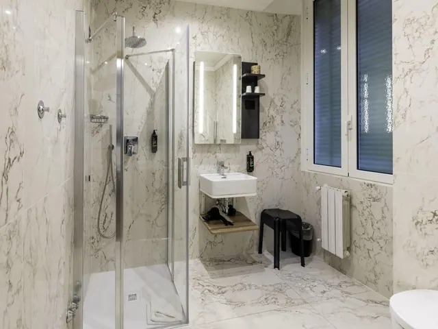 A marble-tiled bathroom with a glass-enclosed walk-in shower, a wall-mounted sink with a lit mirror, a radiator and a window with blue blinds.