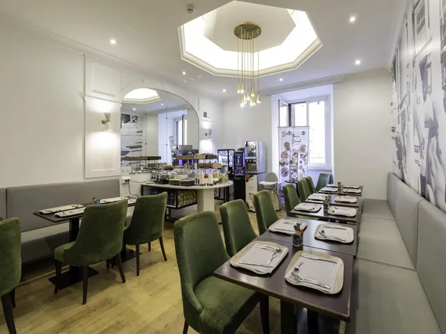 A breakfast room with green velvet chairs at set tables, banquette seating along the walls, and a buffet counter with coffee machines at the back under an octagonal skylight.