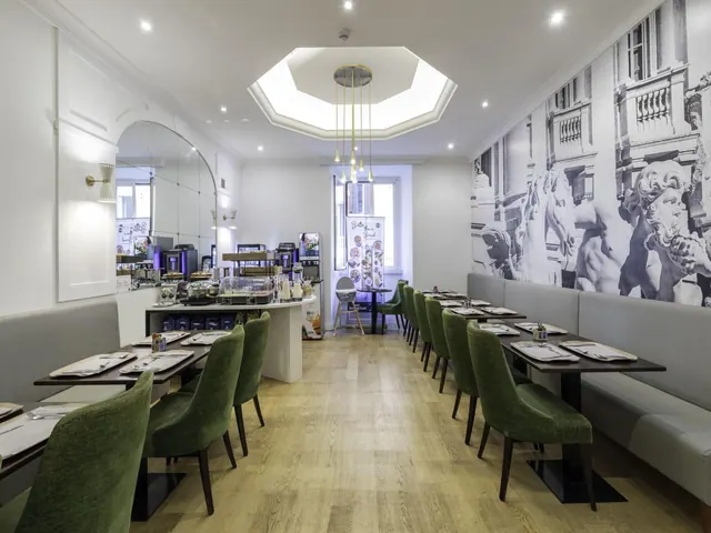 A long dining room with green upholstered chairs, grey banquette seating, a black-and-white statue mural on the right wall, and a breakfast buffet station at the far end.