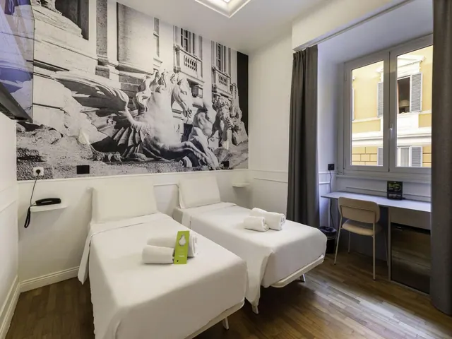 A twin bedroom with two single beds, a black-and-white mural of fountain sculptures on the wall, and a desk and chair beside a window that looks onto a yellow building facade.