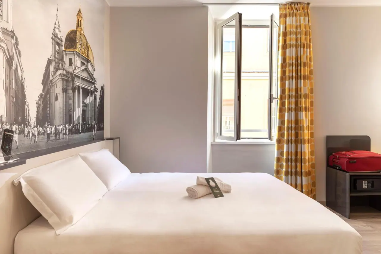 A made double bed with white bedding and folded towels stands in a room with a black-and-white church mural on the wall, an open window with yellow curtains, and a red suitcase on a side table.