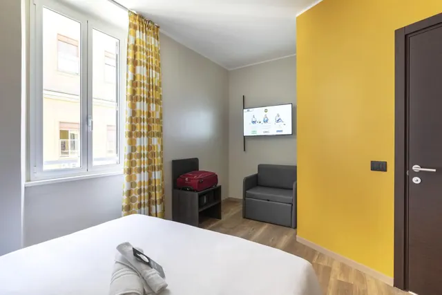 A hotel bedroom with a made bed with white linen and a folded towel, a grey sofa bed, a wall-mounted television, a red suitcase on a dark cabinet, and a yellow accent wall.