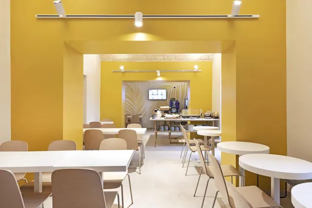 A breakfast dining room with yellow walls, white tables and beige chairs, with a buffet counter and coffee machine at the back.