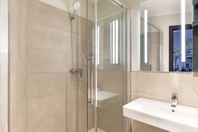 A bathroom with a glass-walled walk-in shower, beige tiled walls, a wall-mounted hand shower, and a white basin with a chrome tap beneath an illuminated mirror.