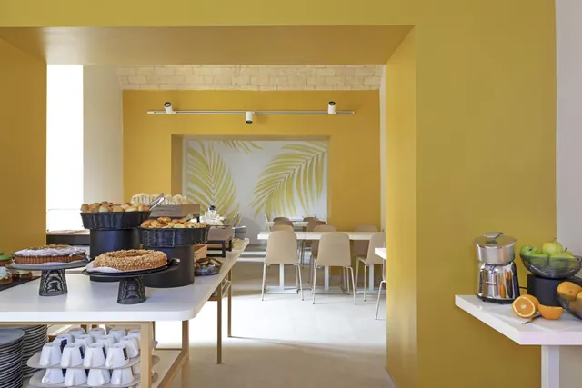 A breakfast buffet with pastries, cakes and stacked white cups on a white counter, with a fruit juicer and fruit bowls beside it, in a yellow dining room with tables and chairs beyond.