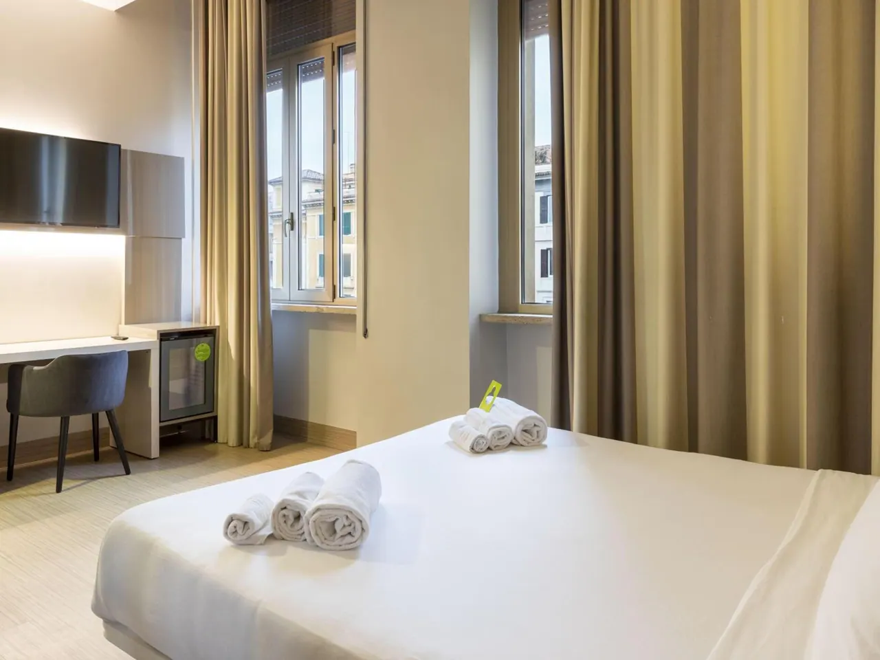 A hotel bedroom with a white bed topped with rolled towels, a desk and chair, a wall-mounted television, a minibar, and two windows looking onto neighbouring buildings.