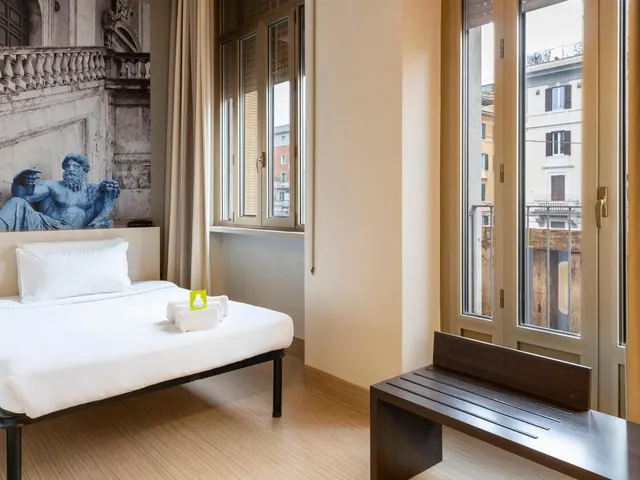 A bedroom with a made bed with white linen and towels, a blue-toned statue mural on the wall, a wooden bench, and tall windows looking onto neighbouring buildings.