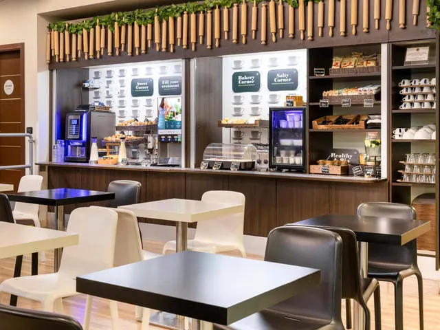 A breakfast area with a self-service counter holding a coffee machine, pastries in baskets, cups on shelves and a glass-door fridge, with empty black tables and white chairs in front.