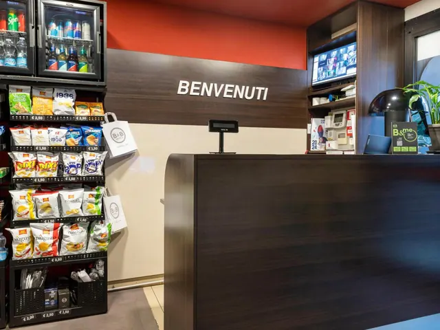 A reception desk with a BENVENUTI sign stands beside a snack and drink display, a wall-mounted TV and a potted plant.