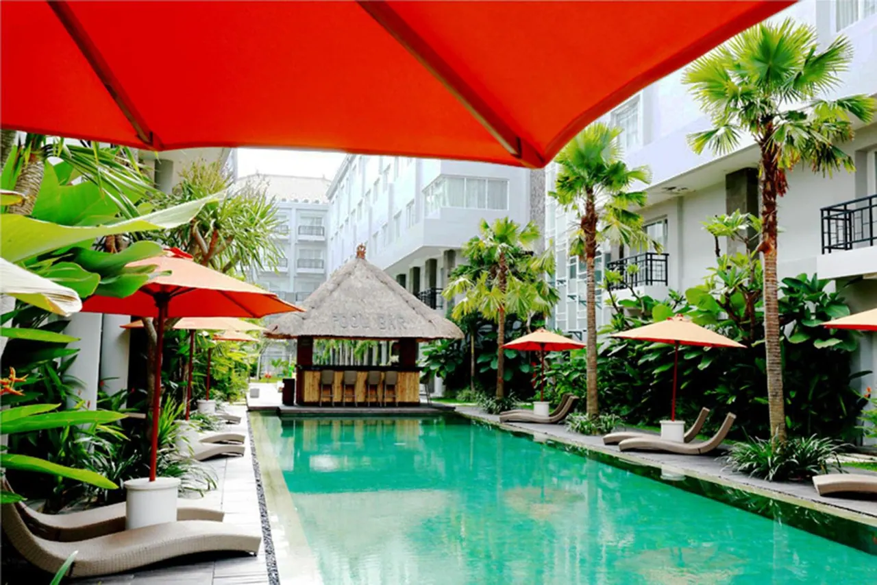 A swimming pool with red umbrellas, sun loungers, palm trees and a thatched-roof pool bar beside hotel buildings.