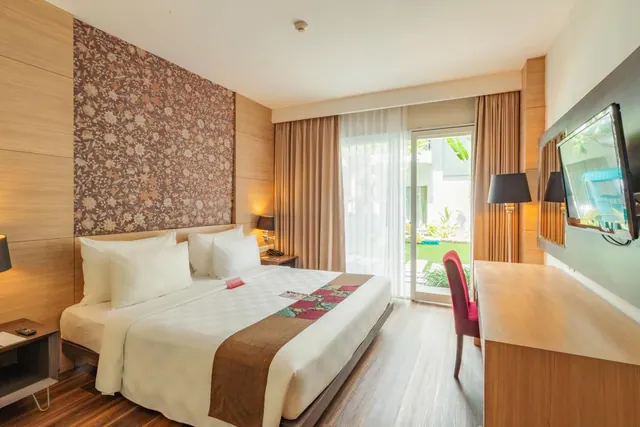A king bed with white linen and a batik-patterned wall faces a wooden desk and wall-mounted television, with a glass door opening onto a garden.
