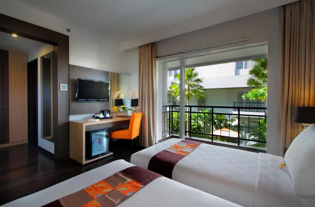 A hotel room with two beds, a wall-mounted television, a desk with an orange chair and a minibar, and sliding doors onto a balcony overlooking palm trees and a neighbouring building.