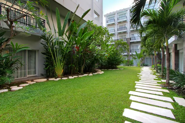 A grassy courtyard garden lined with tropical plants and palm trees, with a white stepping-stone path and hotel buildings behind.