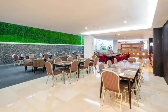 A bright restaurant with rows of wooden tables and upholstered chairs, a green living wall on the left, and red lounge chairs in the background.