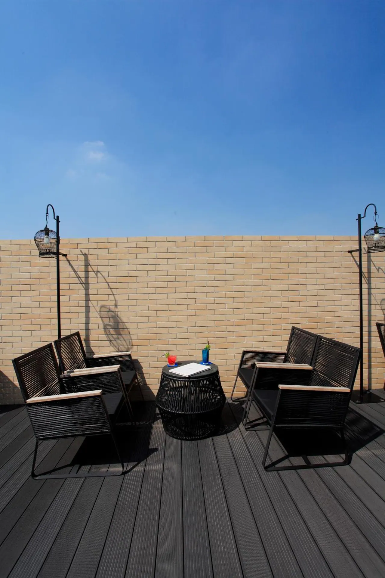 A rooftop terrace with black woven armchairs, a round side table holding drinks and a notepad, lamps on poles, and a brick wall under a clear blue sky.