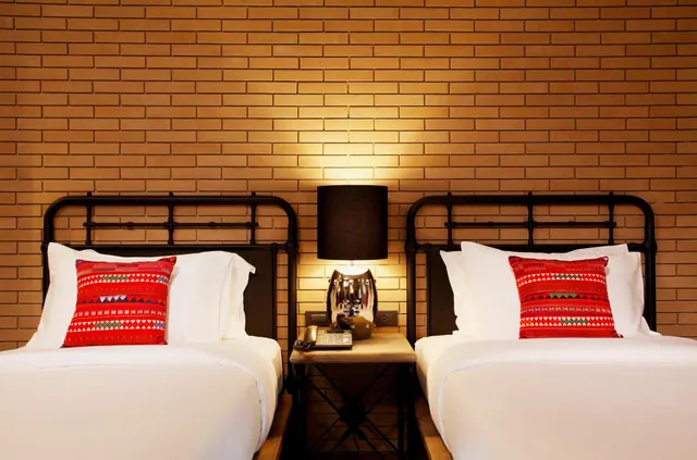 Two single beds with black metal headboards and red patterned cushions sit either side of a bedside table with a lamp against a brick wall.