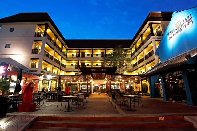 Twilight view of a multi-storey hotel building with lit balconies, courtyard tables and chairs under lit pergolas, and a blue illuminated sign on the right.