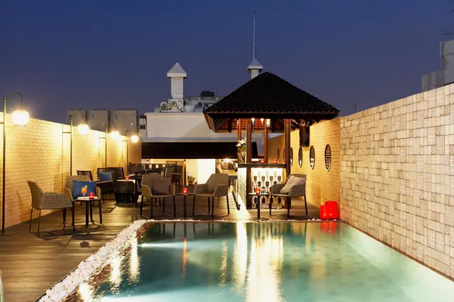 A rooftop pool at dusk beside a wooden deck with outdoor chairs and tables, a covered bar and a tiled wall.