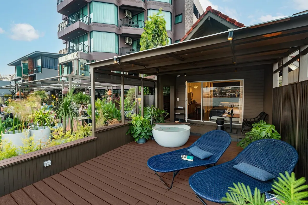 A covered wooden terrace with two blue loungers, a round white soaking tub, potted plants and a glass-walled room behind.