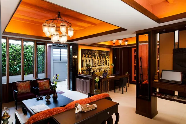 A living area with an orange-lit coffered ceiling, a chandelier, sofas with orange cushions, a dark wood coffee table, a desk and television, and floor-to-ceiling windows looking onto foliage.