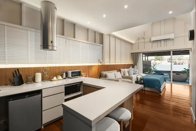 A studio room with an island kitchen counter and stools, a sofa and bed, and sliding doors opening onto a deck with a freestanding tub and a view of boats on the water.