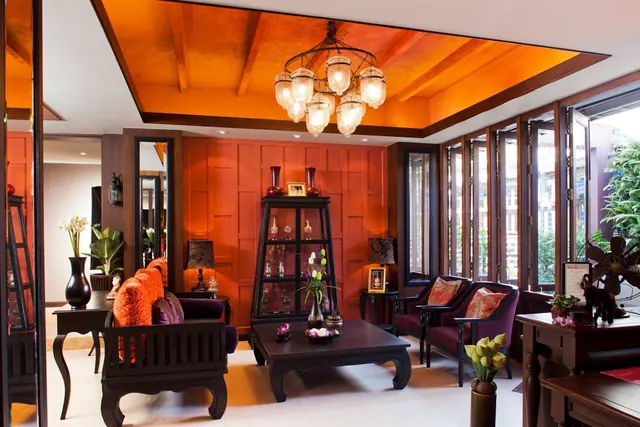A seating area with dark wooden sofas and purple cushions, a glass display cabinet, and an ornate chandelier under an orange ceiling, with folding glass doors looking onto greenery.