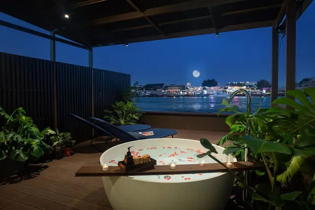 A freestanding bathtub with rose petals and candles sits on an outdoor terrace overlooking a river with lit buildings at night.
