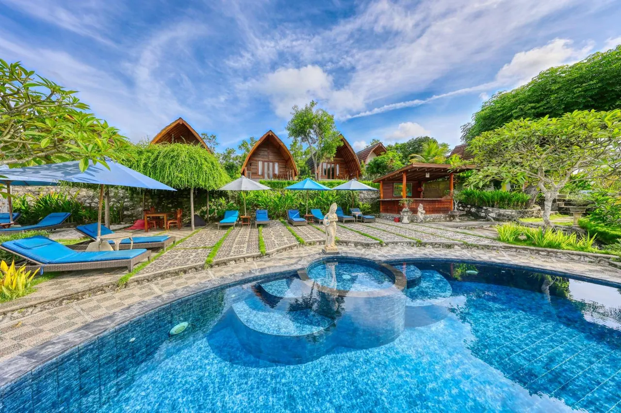 A large curved pool with a circular inset spa, blue sun loungers and umbrellas, and thatched wooden bungalows in the background under a partly cloudy blue sky.