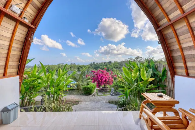 Looking out through an arched wooden doorway from a tiled terrace onto a garden with pink bougainvillea, banana plants and trees under a partly cloudy sky.