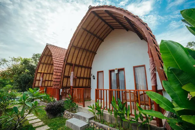A bungalow with a curved thatched roof, white walls, and a wooden balcony railing, surrounded by tropical plants and a stone path.