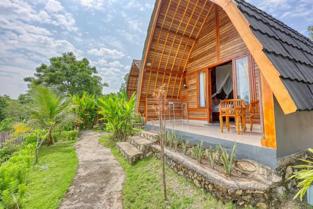 A wooden bungalow with a dark tiled roof and a covered terrace with chairs stands beside a stone-edged garden path and planted beds.