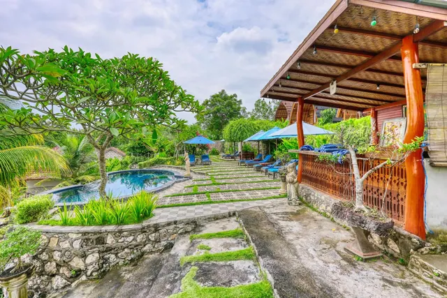 A small kidney-shaped swimming pool with sun loungers and blue parasols sits beside a covered wooden terrace, with frangipani trees and greenery behind.