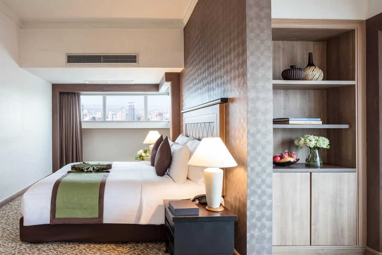 A bedroom with a made king bed, two bedside lamps, open shelving with decorative vases, and a window showing a city skyline.