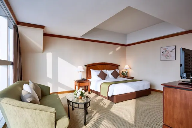 A bedroom with a king bed, a green sofa and a glass coffee table with flowers, a wooden cabinet holding a flat-screen television, and large windows on the left.