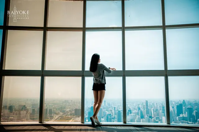 A woman in shorts stands with her back to the camera at floor-to-ceiling windows overlooking a hazy city skyline, with a Baiyoke Sky Hotel logo watermark in the top left.